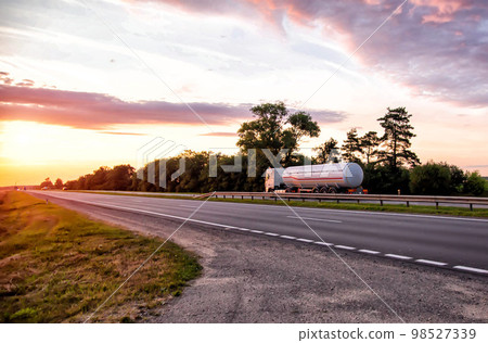 A truck with a semi-trailer tanker transports dangerous goods against the backdrop of a sunny sunset and the sky in the clouds. Transportation of fluid and liquid cargo in trucking industry A truck with a semi-trailer tanker transports dangerous goods against the backdrop of a sunny sunset and the sky in the clouds. Transportation of fluid and liquid cargo in trucking industry 98527339