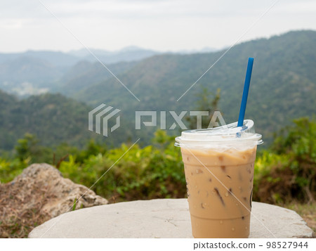 A glass of iced coffee is placed on a rock against a background of mountains and sky. A glass of iced coffee is placed on a rock against a background of mountains and sky. 98527944