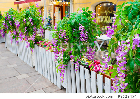 Outdoor cafe on street decorated artificial  purple flowers Ivy wisteria, green plant, white fence. Cafe summer terrace. Text translation: author's cuisine. 98528454