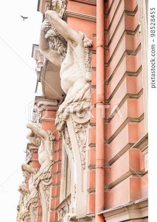 Atlant, Atlas or Titan sculpture on pink historic building and bird in sky. Male man sculpture with beard, full length, side view, vertical photo. Atlant, Atlas or Titan sculpture on pink historic building and bird in sky. Male man sculpture with beard, full length, side view, vertical photo. 98528455