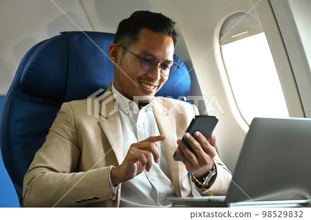 Millennial businessman passenger sitting on airplane cabin and using smart phone. Business or travel abroad concept Millennial businessman passenger sitting on airplane cabin and using smart phone. Business or travel abroad concept 98529832