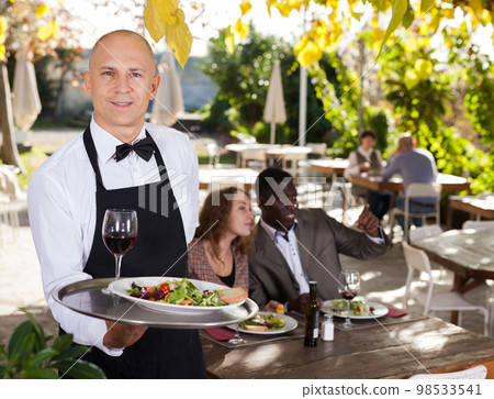 Adult waiter serves customers in a open-air restaurant Adult waiter serves customers in a open-air restaurant 98533541