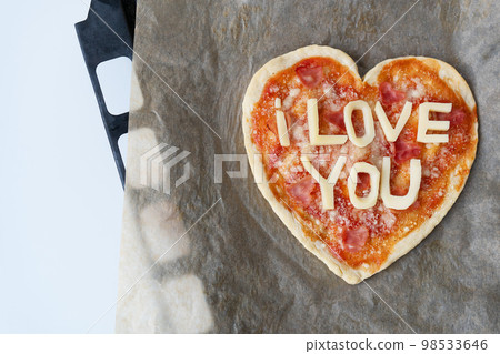 Italian heart shaped pizza with salami, tomato sauce, parmesan, pizza sauce, mozzarella and olive oil on parchment paper with cheese I love you. Love concept for Valentine's Day February 14. Italian heart shaped pizza with salami, tomato sauce, parmesan, pizza sauce, mozzarella and olive oil on parchment paper with cheese I love you. Love concept for Valentine's Day February 14. 98533646