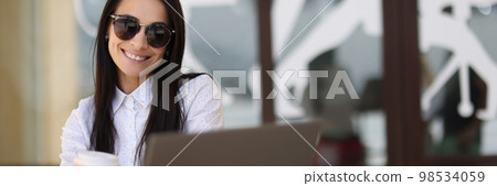 Smiling sun woman holding coffee and working on laptop in cafe Smiling sun woman holding coffee and working on laptop in cafe 98534059