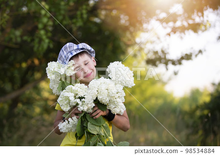 Cheerful happy village child with a bouquet of flowers. Smiling little boy with hydrangea in the garden. Mother's day, March 8, International Women's Day concept. Cheerful happy village child with a bouquet of flowers. Smiling little boy with hydrangea in the garden. Mother's day, March 8, International Women's Day concept. 98534884