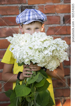 Cheerful happy village child with a bouquet of flowers. Smiling little boy with hydrangea near the grunge brick wall. Mother's day, March 8, International Women's Day concept. Cheerful happy village child with a bouquet of flowers. Smiling little boy with hydrangea near the grunge brick wall. Mother's day, March 8, International Women's Day concept. 98534885