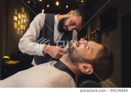 Professional barber carefully trimming the clients beard Professional barber carefully trimming the clients beard 98536664
