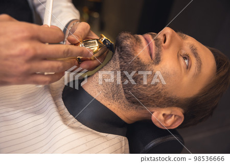 Professional barber carefully trimming the clients beard Professional barber carefully trimming the clients beard 98536666