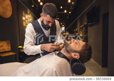 Professional barber carefully trimming the clients beard Professional barber carefully trimming the clients beard 98536669