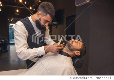 Professional barber carefully trimming the clients beard Professional barber carefully trimming the clients beard 98536671