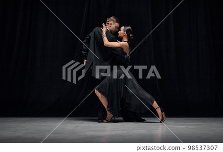 Passionate dance style. Man and woman, professional tango dancers performing in black stage costumes over black background. Passionate dance style. Man and woman, professional tango dancers performing in black stage costumes over black background. 98537307