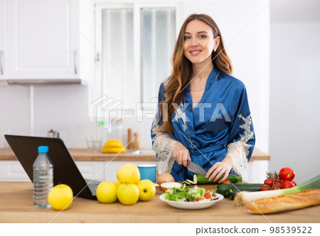 Young woman in blue robe cooking and watching TV series on laptop 98539522
