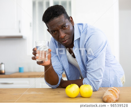 African american tired man drinking water after preparing vegetable salad in kitchen 98539528