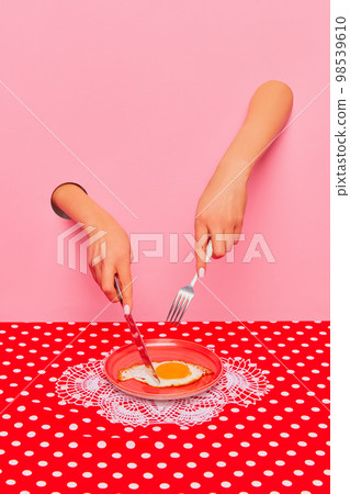 Food pop art photography. Female hand sticking out pink paper and cutting fried eggs on plate on retro tablecloth. Breakfast time 98539610