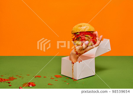 Food pop art photography. Female hand sticking out food box with burger on green tablecloth and orange background Food pop art photography. Female hand sticking out food box with burger on green tablecloth and orange background 98539654