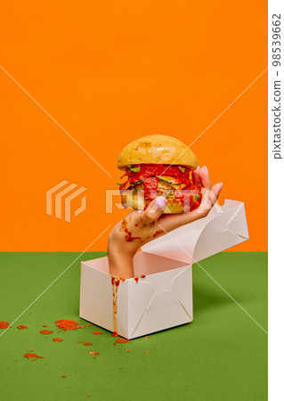Food pop art photography. Female hand sticking out food box with delicious burger on green tablecloth and orange background 98539662