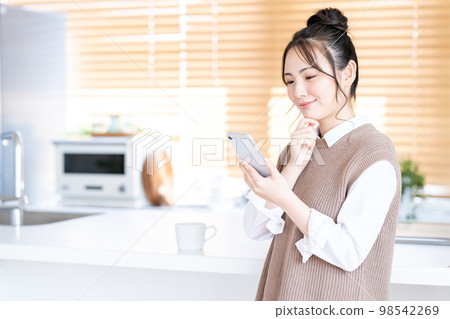 A young woman operating a smartphone in the kitchen. 98542269