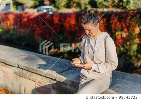 Young Fashionable Teenage Girl With Smartphone In Europian Park In Autumn Sitting At Smiling. Trendy Young Woman In Fall In Park Texting. Retouched, Vibrant Colors. Beautiful Blonde Teenage Girl Young Fashionable Teenage Girl With Smartphone In Europian Park In Autumn Sitting At Smiling. Trendy Young Woman In Fall In Park Texting. Retouched, Vibrant Colors. Beautiful Blonde Teenage Girl 98543722