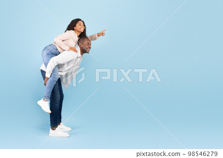 Happy black couple having fun, lady piggybacking man and pointing finger at free space on blue background, banner Happy black couple having fun, lady piggybacking man and pointing finger at free space on blue background, banner 98546279
