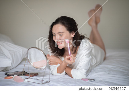 Cheerful young european lady in bathrobe lies on white bed, apply cosmetics with brush on face, looks in mirror 98546327