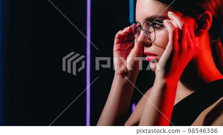 Retro optic. 90s look. 2000s eyewear fashion. Red purple color neon light closeup portrait of teen girl model face wearing round glasses on dark black copy space background. Retro optic. 90s look. 2000s eyewear fashion. Red purple color neon light closeup portrait of teen girl model face wearing round glasses on dark black copy space background. 98546386