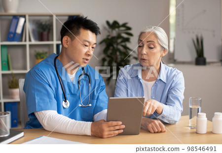 Serious middle aged korean male doctor showing tablet and consulting senior european lady patient in clinic 98546491