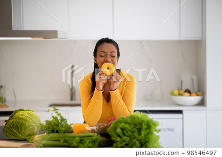 Cheerful hungry millennial african american lady in casual hold donut, enjoy aroma at table with organic vegetables 98546497