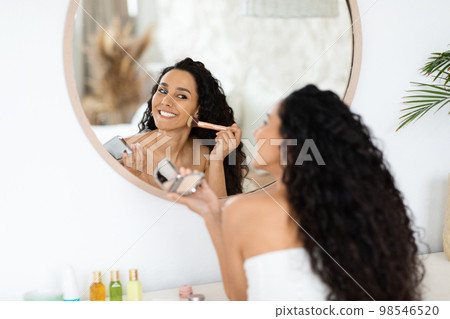 Smiling pretty arab millennial curly lady in towel applying blush on face, looking in mirror, enjoy daily procedure Smiling pretty arab millennial curly lady in towel applying blush on face, looking in mirror, enjoy daily procedure 98546520