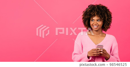 Cheerful millennial african american curly lady with braces in casual typing on phone looks at copy space 98546525