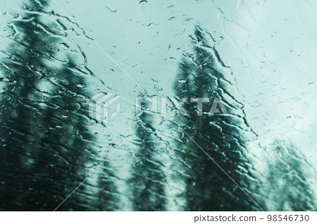 Drops on car glass against mountain forest. Droplets fall on window glass against thick coniferous trees in forest Drops on car glass against mountain forest. Droplets fall on window glass against thick coniferous trees in forest 98546730