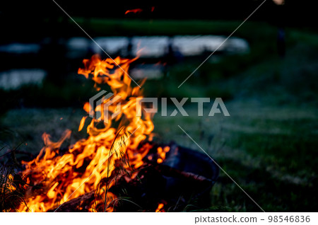 Leaping flames of a camp fire with a darkening view of a pond in the background. 98546836