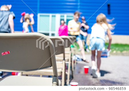 Selective focus on rows of metal folding chairs at an outdoor school function with blurred children in the background. Selective focus on rows of metal folding chairs at an outdoor school function with blurred children in the background. 98546859