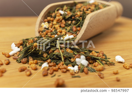 Green tea Japanese Genmaicha. Green tea mixed with roasted popped brown rice in wooden scoop on bamboo board. brown background behind. macro photo. close up. shallow depth of field. 98547997
