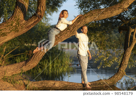 Latin woman lying on green tree branch, Caucasian man stands nearby and look into distance, happy couple enjoys date, time together, having fun near water. Lovers wear casual clothes. Horizontal plane Latin woman lying on green tree branch, Caucasian man stands nearby and look into distance, happy couple enjoys date, time together, having fun near water. Lovers wear casual clothes. Horizontal plane 98548179