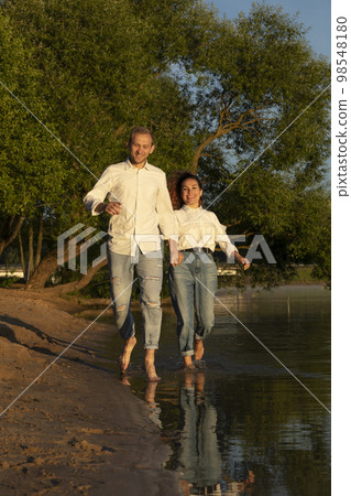 Caucasian man runs holding Latin woman hand on beach, happy couple enjoys time together, having fun near water. Green trees on background. Lovers wear casual clothes. Vertical plane Caucasian man runs holding Latin woman hand on beach, happy couple enjoys time together, having fun near water. Green trees on background. Lovers wear casual clothes. Vertical plane 98548180