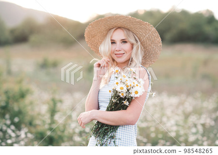 Smiling beautiful blonde woman 25-29 year old holding bouquet of fresh daisy wearing straw hat and dress posing in meadow close up. Smiling beautiful blonde woman 25-29 year old holding bouquet of fresh daisy wearing straw hat and dress posing in meadow close up. 98548265