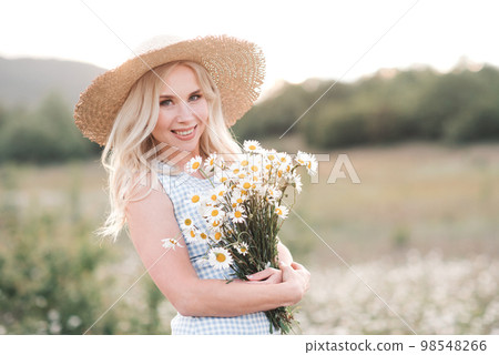 Smiling beautiful blonde woman 25-29 year old holding bouquet of fresh daisy wearing straw hat and dress posing in meadow close up. Smiling beautiful blonde woman 25-29 year old holding bouquet of fresh daisy wearing straw hat and dress posing in meadow close up. 98548266