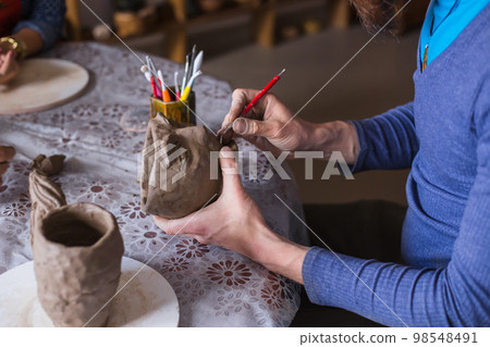 Professional potter carving mug with special tool in pottery workshop 98548491