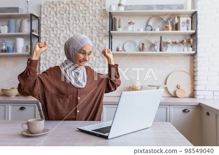 Freelancer in hijab working on laptop remotely from home, woman received good news and good news online notification, happy and celebrating good achievement results, businesswoman in home office. Freelancer in hijab working on laptop remotely from home, woman received good news and good news online notification, happy and celebrating good achievement results, businesswoman in home office. 98549040