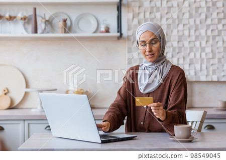 Cheerful and successful woman in hijab at home in kitchen with laptop and bank credit card makes online purchase in online store, Muslim woman chooses products and gifts remotely. Cheerful and successful woman in hijab at home in kitchen with laptop and bank credit card makes online purchase in online store, Muslim woman chooses products and gifts remotely. 98549045