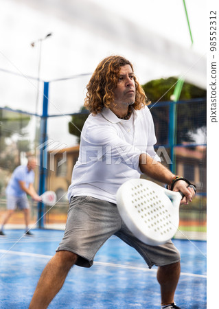Focused middle-aged Latin man playing padel on court. View through tennis net Focused middle-aged Latin man playing padel on court. View through tennis net 98552312