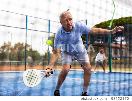 Positive elderly male player serving ball during training padel in court. View through tennis net 98552370