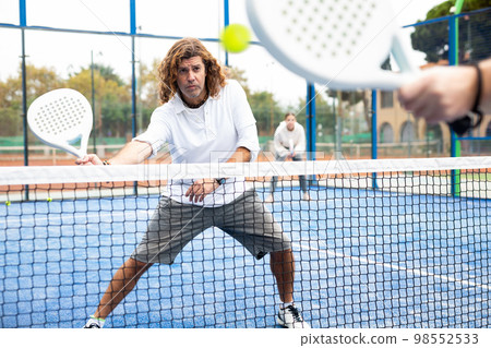 Focused middle-aged Latin man padel player hitting ball with racket on hard court in summer Focused middle-aged Latin man padel player hitting ball with racket on hard court in summer 98552533