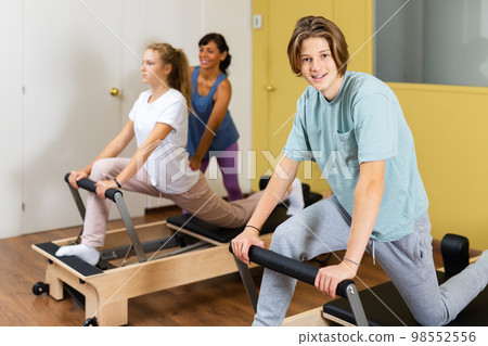 Young boy looking in camera while training pilates in gym. Their trainer hispanic woman helping girl with exercises on pilates reformer in background. 98552556