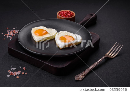 Two heart-shaped fried eggs on a black ceramic plate on a dark concrete background 98559956