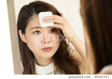 A woman is looking in the mirror and wiping her forehead with a cotton pad. 98561417