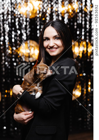 Young beautiful smiling brunette female in black jacket holding her litlle dog in her hands. Carefree woman posing near shiny tinsel wall in studio with dog. 98563692