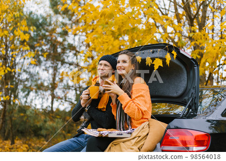 A happy young family is relaxing after a day spent outdoors in the autumn forest. A couple in love is sitting in the trunk of a car and drinking tea from a thermos with their dog. Travel in the autumn 98564018