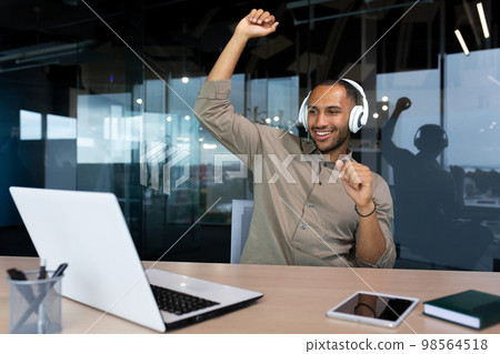 Happy young African American man sitting in office at desk with laptop wearing white headphones. He listens to music, dances, raised his hands up, rests, a break. Happy young African American man sitting in office at desk with laptop wearing white headphones. He listens to music, dances, raised his hands up, rests, a break. 98564518