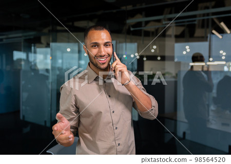 A young African American man is standing in the office and talking on the phone. gesturing with his hands, looking at the camera, smiling. A young African American man is standing in the office and talking on the phone. gesturing with his hands, looking at the camera, smiling. 98564520